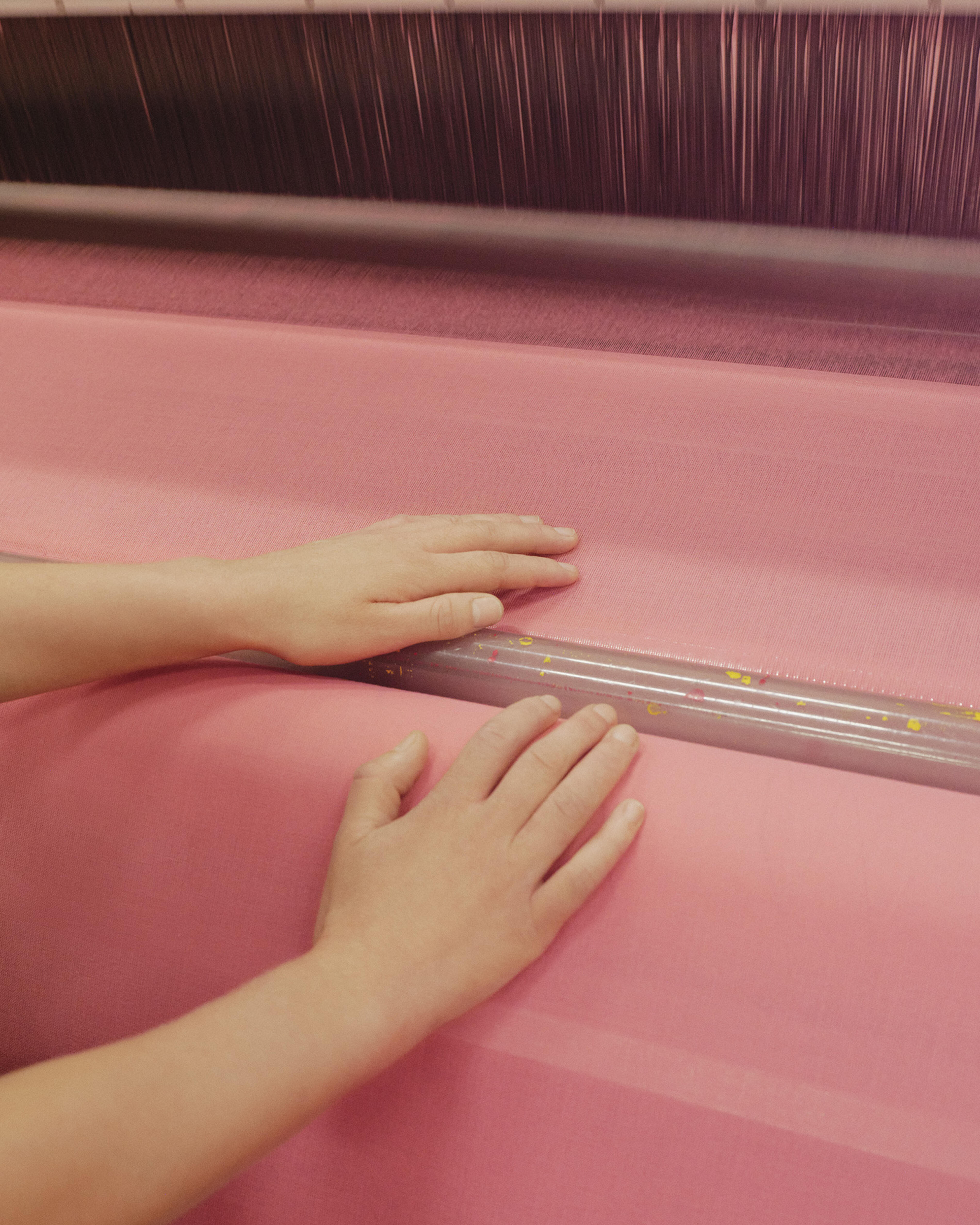 Two hands work on a loom making a pink fabric