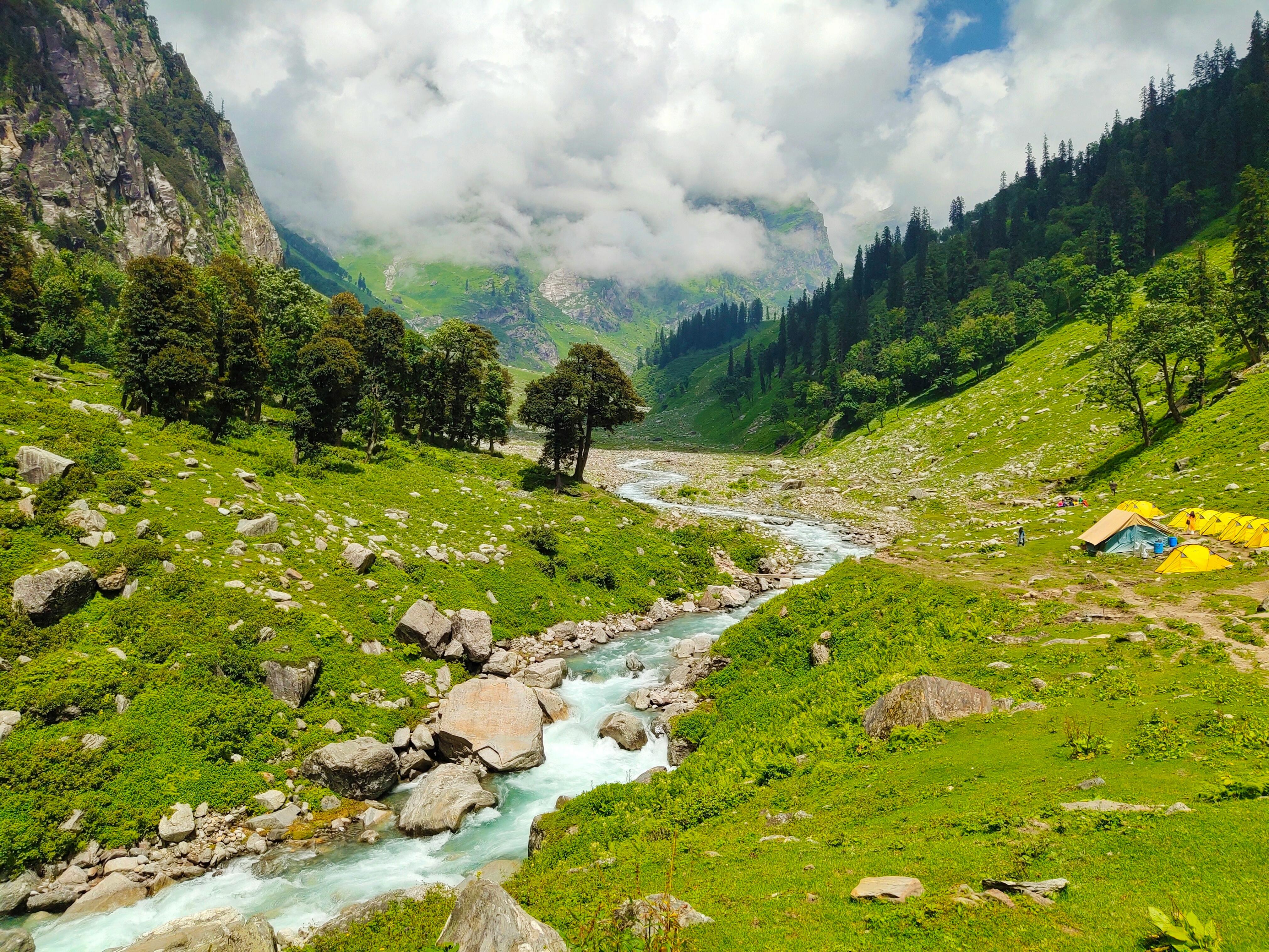 Mountain landscape of the Himalayas, with a stream running down a valley of green lush vegetation and mountains in the background.