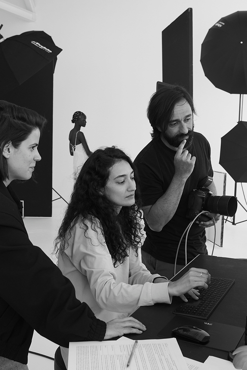 A photographer and two other people from the ecommerce web team check an image on the computer while the model awaits in the set behind them.