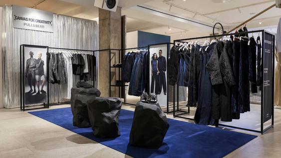 Pull&Bear fashion display with dark and blue clothes on shelves, mannequins and pedestals on a blue carpet.