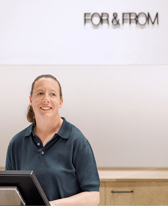 Smiling woman in dark polo shirt behind a counter with a "FOR & FROM" sign in the background.