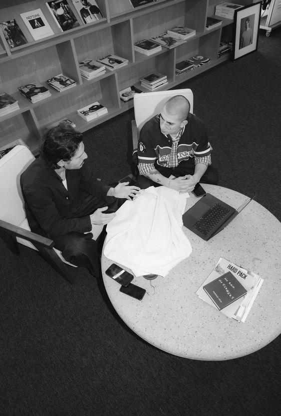 Two men sitting at a round table in a bookstore, talking with a laptop, phones and magazines on the table.