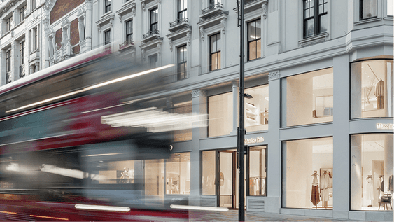 A red London bus passes in front of a Massimo Dutti store located in a modern city building.