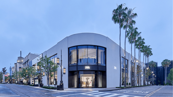 Modern Zara store with curved facade and large windows, surrounded by palm trees at dusk.