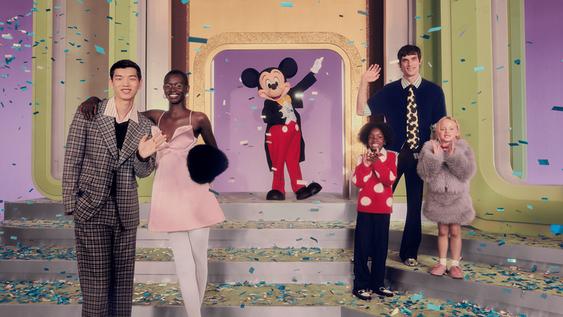 A group of adult and child models pose next to Mickey Mouse in a colorful and festive setting with confetti.