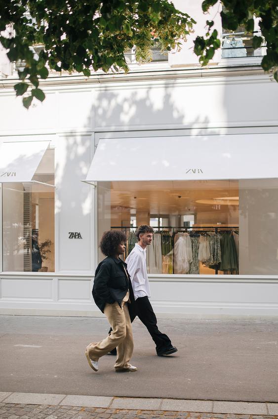 Two people walking along a tree-lined street in front of a Zara store.